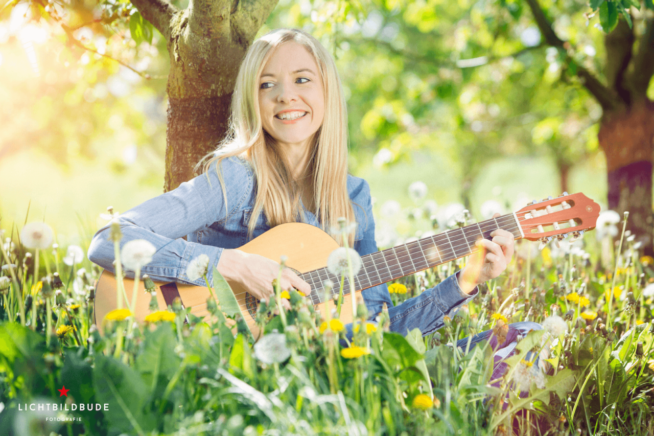 Nadine Petry sitzt mit einer Gitarre unter einem Baum in einer Blumenwiese